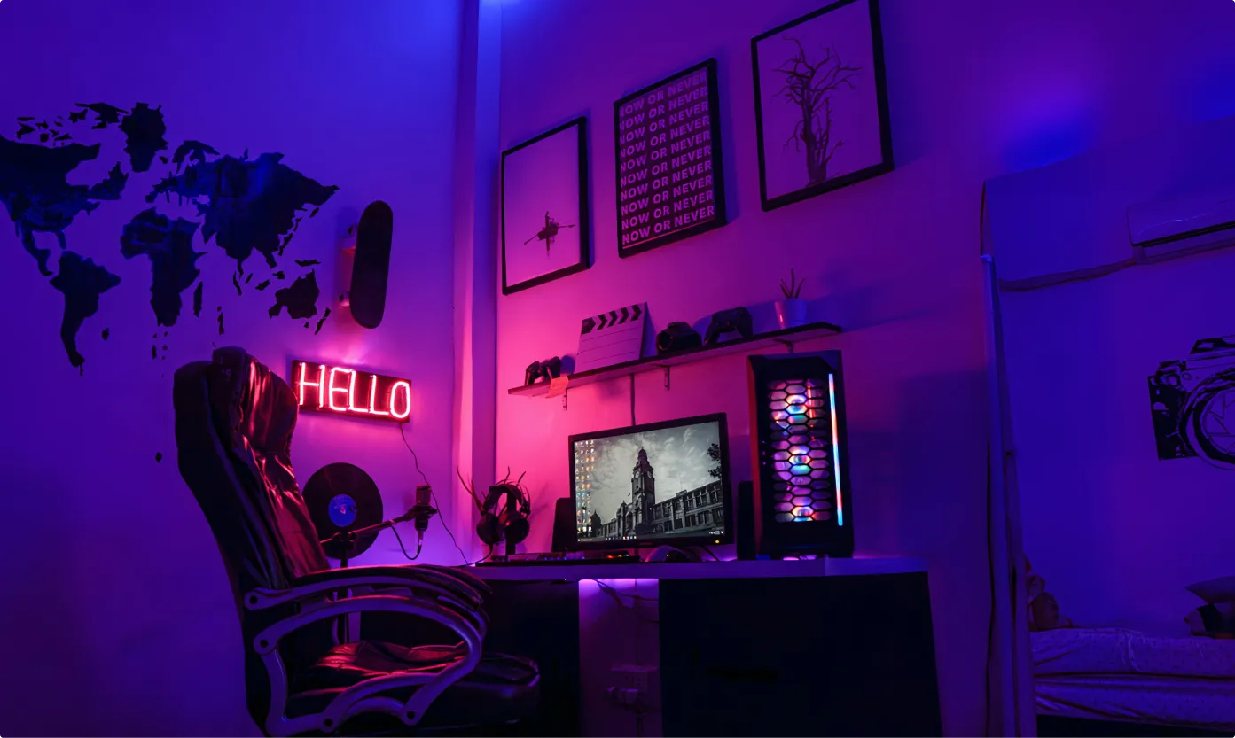 Gaming setup with a black leather chair, a desk holding a computer monitor and a colorful PC tower, illuminated by purple and pink neon lights with wall decorations including a 'HELLO' neon sign and framed art.